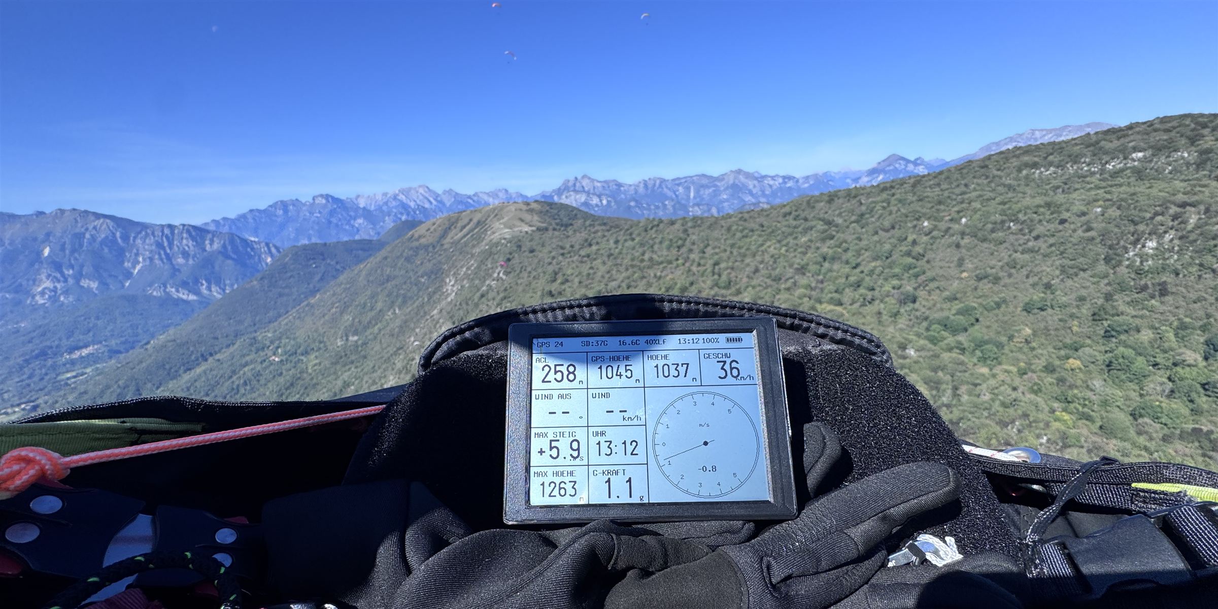 Blick vom Gleitschirm im Flug: fliteai-Vario auf dem Cockpit, dahinter Bergkette mit weiteren Gleitschirmen am Himmel